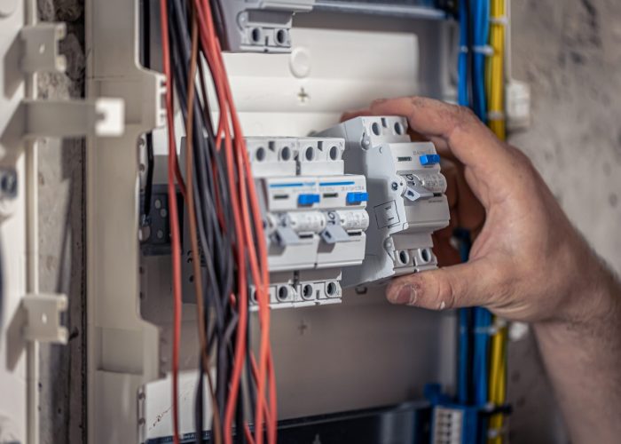 A male electrician works in a switchboard with an electrical connecting cable, connects the equipment with tools.